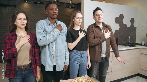Multi ethnic group of friends listening and singing national anthem standing and hand on heart before watching football match on TV together at home