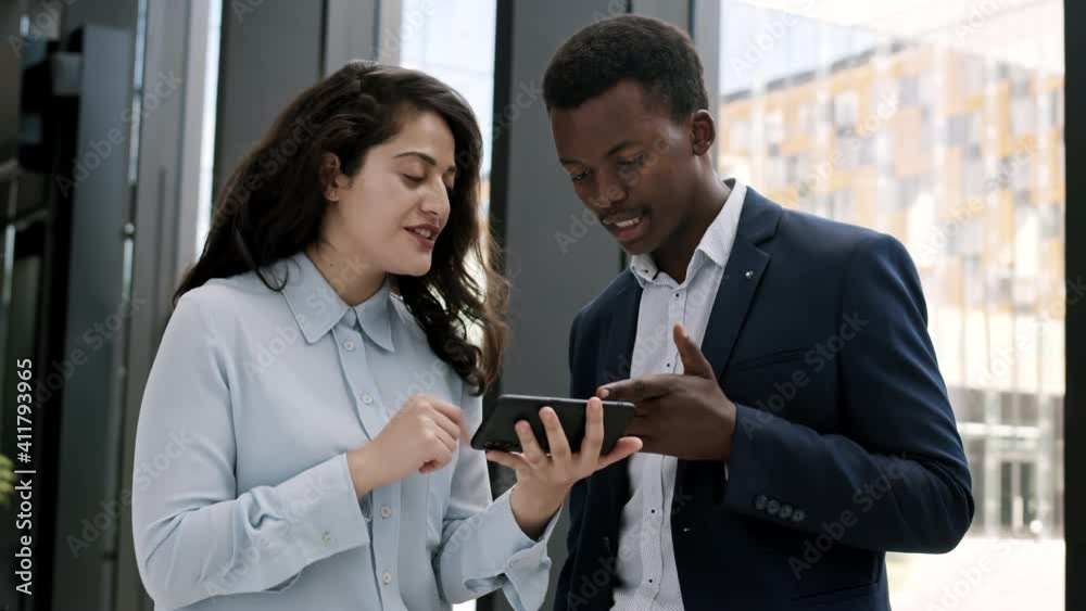 Medium shot of African-American businessman and his female mixed-race colleague standing together in office hall and looking at telephone screen woman holding in hands