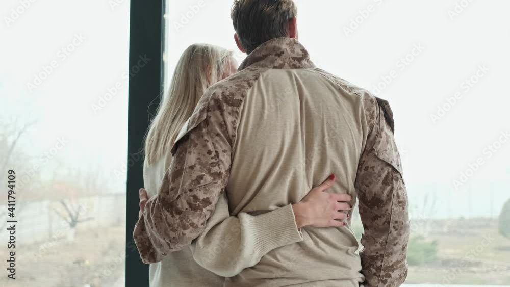 A back view of calm couple young american soldier man and woman are hugging each other while looking to the window standing inside the apartments A back view of calm couple young american soldier man and woman are hugging each other while looking to the window standing inside the apartments
