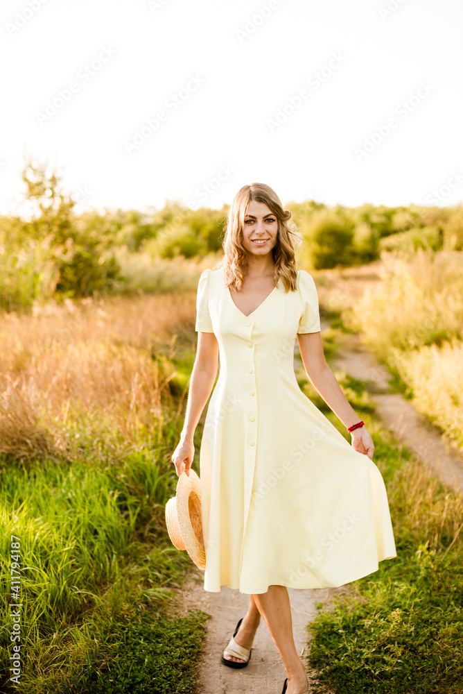 A young beautiful girl with blond curly hair in a light summer yellow dress and with a straw hat in her hands in a field in the countryside at sunset. People and nature A young beautiful girl with blond curly hair in a light summer yellow dress and with a straw hat in her hands in a field in the countryside at sunset. People and nature