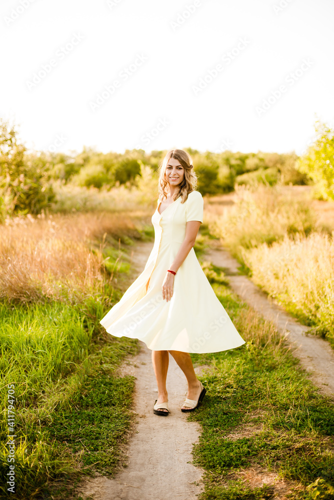 A young beautiful girl with blond curly hair in a light summer yellow dress and with a straw hat in her hands in a field in the countryside at sunset. People and nature A young beautiful girl with blond curly hair in a light summer yellow dress and with a straw hat in her hands in a field in the countryside at sunset. People and nature