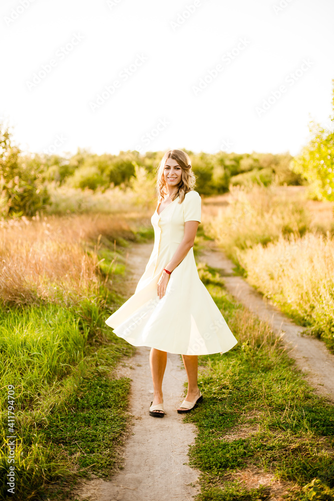 A young beautiful girl with blond curly hair in a light summer yellow dress and with a straw hat in her hands in a field in the countryside at sunset. People and nature A young beautiful girl with blond curly hair in a light summer yellow dress and with a straw hat in her hands in a field in the countryside at sunset. People and nature
