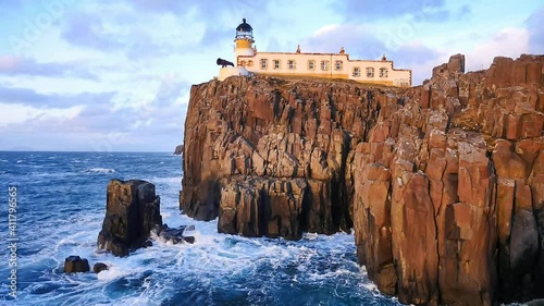 Neist Point lighthouse on west coast of the Isle of Skye in Scotland during an stormy sunset. Shinning lighthouse stand above sea of the Hebrides, foamy waves crashing against to rock and cliff. 