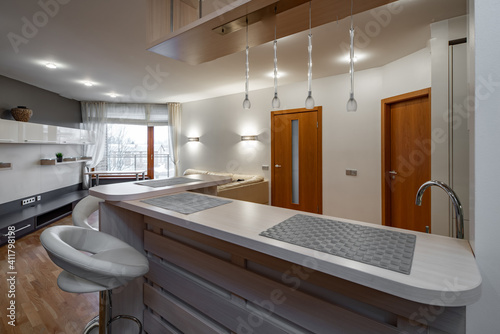 Modern interior of kitchen in luxury flat. New kitchen set with light wooden counter. Bar stool. Living zone with couch. Wooden shelf with drawers.