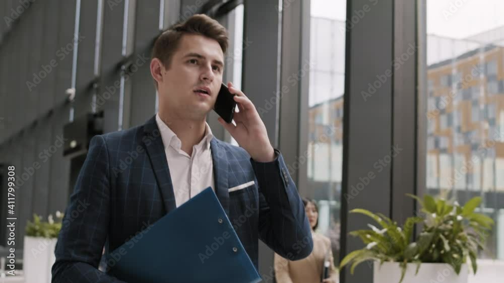 Medium shot of young good-looking Caucasian businessman wearing blue checkered suit going along office hall and having telephone chat with coworker Medium shot of young good-looking Caucasian businessman wearing blue checkered suit going along office hall and having telephone chat with coworker