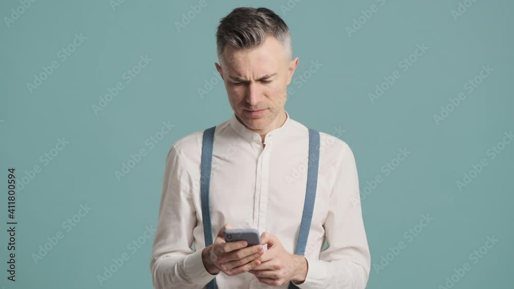 A serious man is using his smartphone standing isolated over blue background wall A serious man is using his smartphone standing isolated over blue background wall