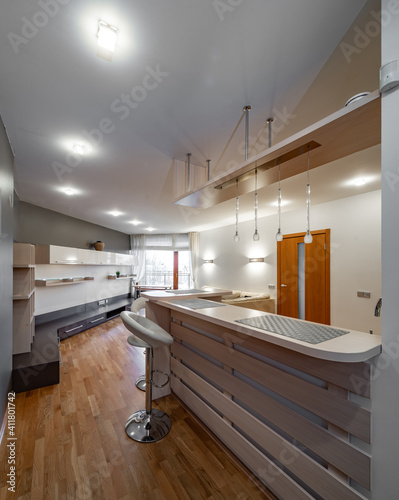 Contemporary interior of kitchen and living room in luxury flat. New kitchen set with light wooden counter. Wooden doors. Bar stool.