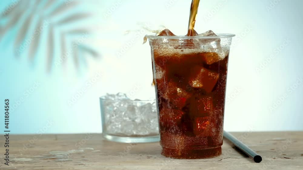 fresh cola drink poring in plastic take away cup with ice cubes forming foams and splashes, defocused palm leaf in background. slow motion