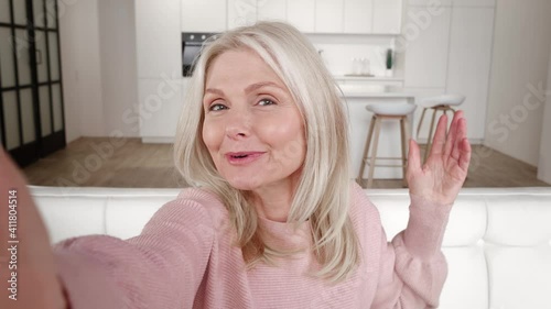 Cheerful senior woman on selfie video conference saying hello facing camera smiling, sitting on sofa at home. Smiling mature lady grandmother holding smartphone do online video call on couch at home.