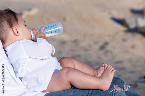 Newborn drinking milk from a baby bottle, sitting on mom legs outdoors