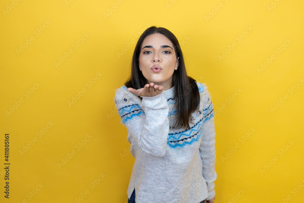 Young caucasian woman wearing casual sweater over yellow background looking at the camera blowing a kiss with hand on air being lovely and sexy. Love expression. Young caucasian woman wearing casual sweater over yellow background looking at the camera blowing a kiss with hand on air being lovely and sexy. Love expression.