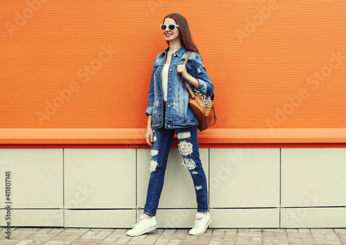 Portrait of smiling young woman wearing a denim jacket and jeans with backpack on a orange background