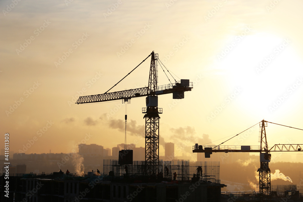 Silhouettes of construction cranes and unfinished residential building on sunrise background. Housing construction, apartment block in winter city
