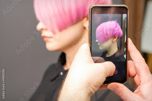 The hairdresser takes pictures of the short pink hairstyle of a young woman on the smartphone
