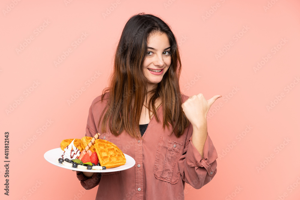 Young caucasian woman holding waffles isolated on pink background pointing to the side to present a product