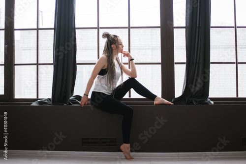 A girl with long pigtails sits on a windowsill near a large window.