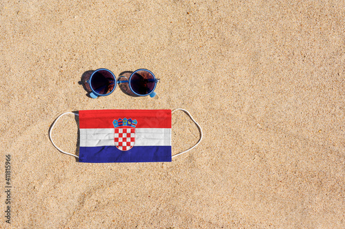 Fototapeta Naklejka Na Ścianę i Meble -  A medical mask in the color of the Croatia flag lies on the sandy beach next to the glasses. The concept of organizing a safe beach holiday in Croatia during the coronavirus pandemic.