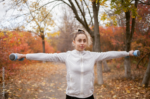 Attractive fitness woman exercising muscles with dumbbells in autumn park