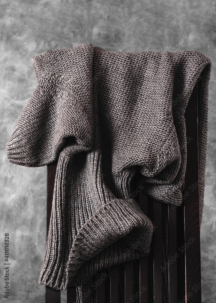 Women's wool knitted gray sweater casually thrown over the back of wooden chair against gray wall. Closeup. Vertical orientation.