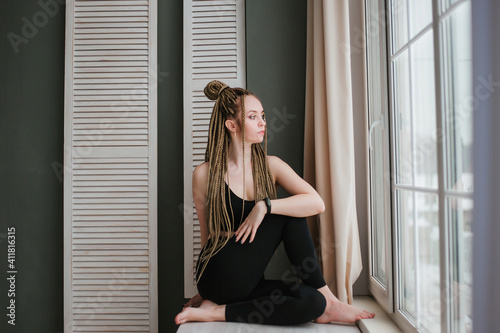 Young woman with long braids in sportswear practices yoga. A beautiful girl is sitting near a large window.