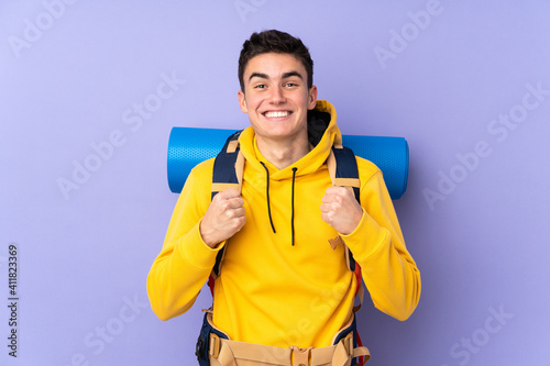 Teenager caucasian mountaineer man with a big backpack isolated on purple background celebrating a victory