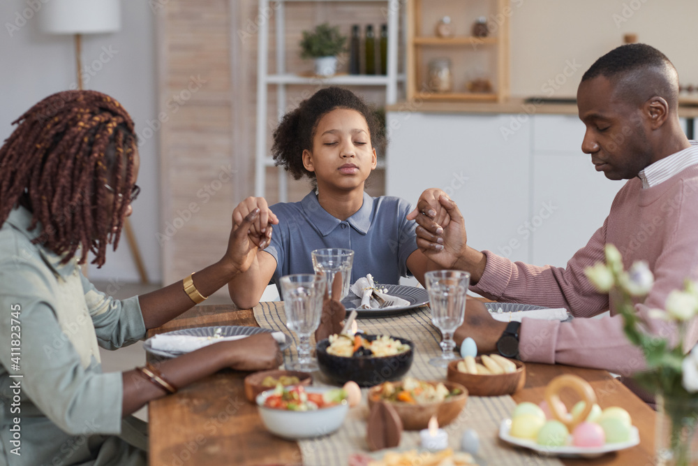 Portrait of modern African-American family saying grace at dinner table on Easter and holding hands, copy space