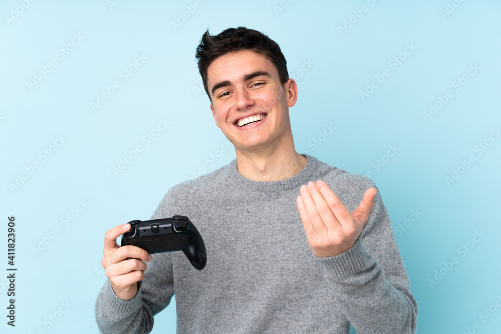 Teenager caucasian man playing with a video game controller isolated on blue background inviting to come with hand. Happy that you came Teenager caucasian man playing with a video game controller isolated on blue background inviting to come with hand. Happy that you came