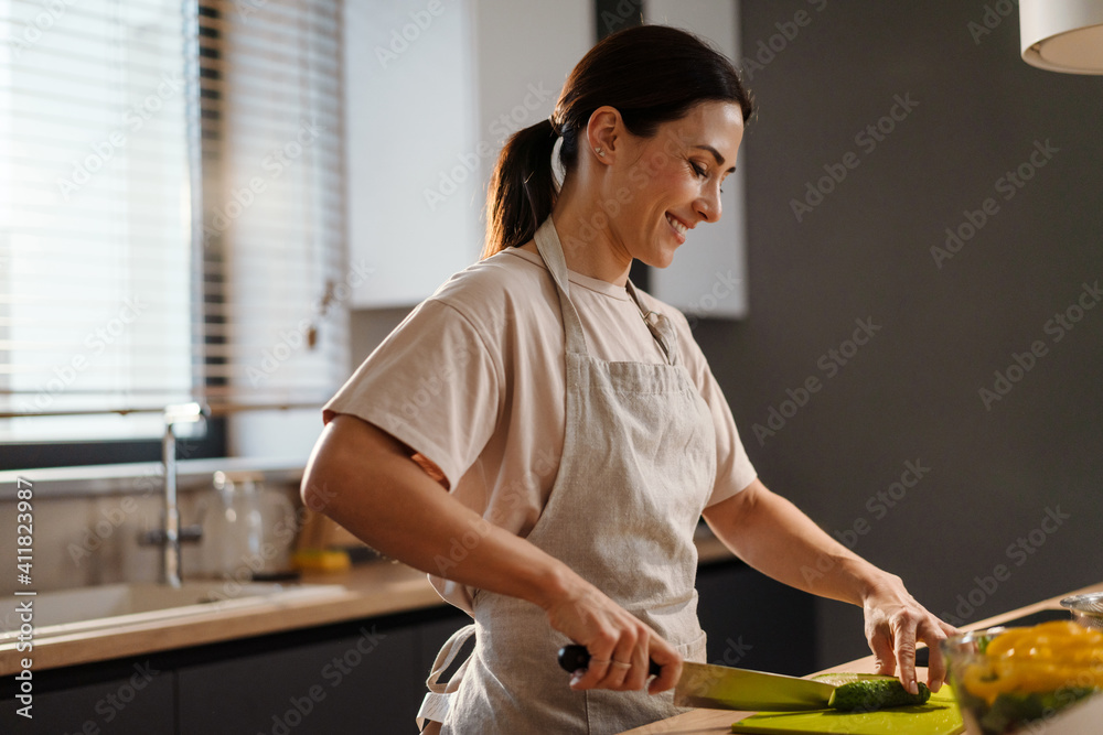 Obraz premium Smiling charming woman making salad with vegetables at home kitchen