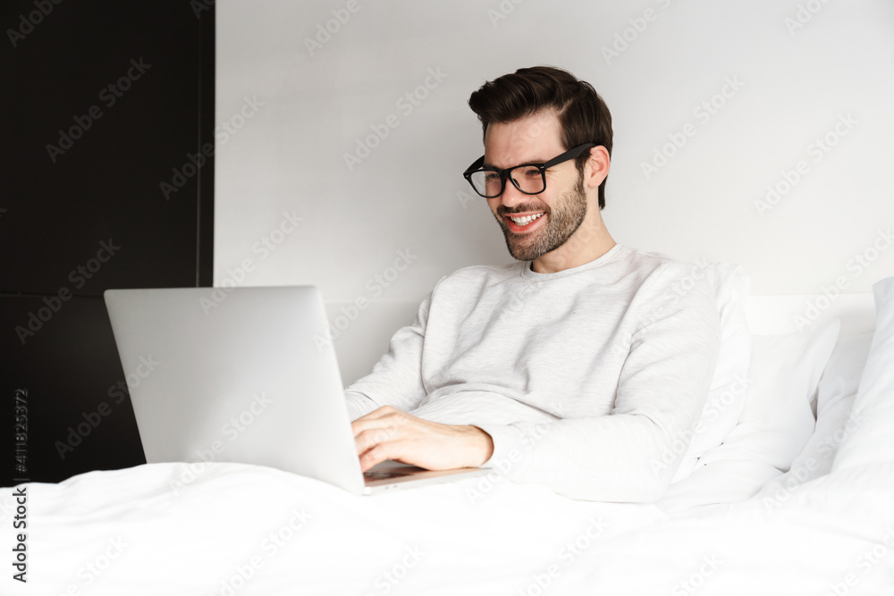 Smiling young man using laptop while resting in bed at home Smiling young man using laptop while resting in bed at home