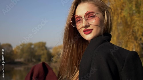 A smiling chic woman in a drape coat and pink-tinted sunglasses takes off her burgundy scarf and throws it gently at the camera, blocking the light of the lens