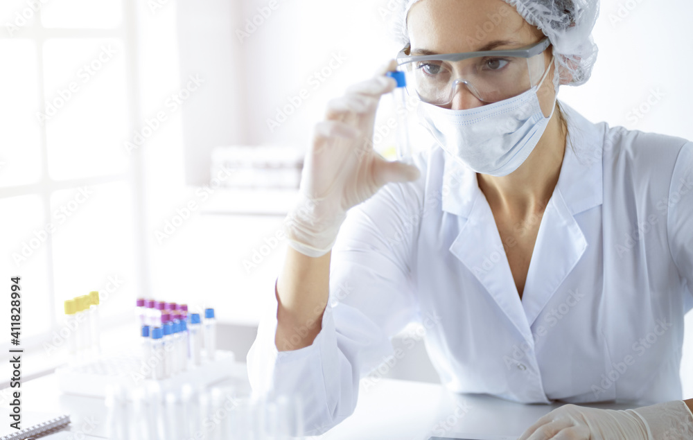 Professional female scientist in protective eyeglasses researching tube with reagents in sunny laboratory. Medicine and science researching Professional female scientist in protective eyeglasses researching tube with reagents in sunny laboratory. Medicine and science researching