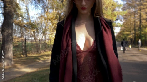 An exhibitionist girl in lace bodysuit lingerie with see through boobs and an open black coat walks down the street among people