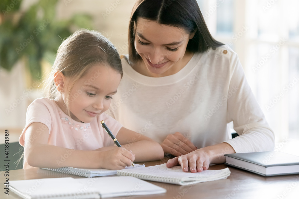 Smiling young mother and little 7s daughter study at home write make homework together. Happy Caucasian mom and small girl child learn distant handwrite prepare school home task or assignment. Smiling young mother and little 7s daughter study at home write make homework together. Happy Caucasian mom and small girl child learn distant handwrite prepare school home task or assignment.