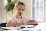 Pensive small cute Caucasian 7s girl sit at desk at home do homework alone write in notebook thinking. Small thoughtful focused little child study learn distant, prepare for school. Education concept.