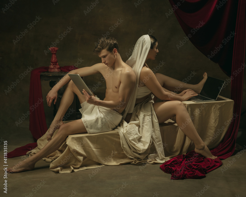 Fototapeta premium Using laptop and tablet. Modern remake of classical artwork with modern tech theme - young medieval couple on dark background, golden colored. Concept of technologies, devices, communication, ad.