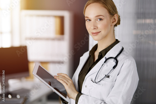 Cheerful smiling female doctor using tablet computer in sunny clinic. Portrait of friendly physician woman at work. Perfect medical service in hospital. Medicine concept