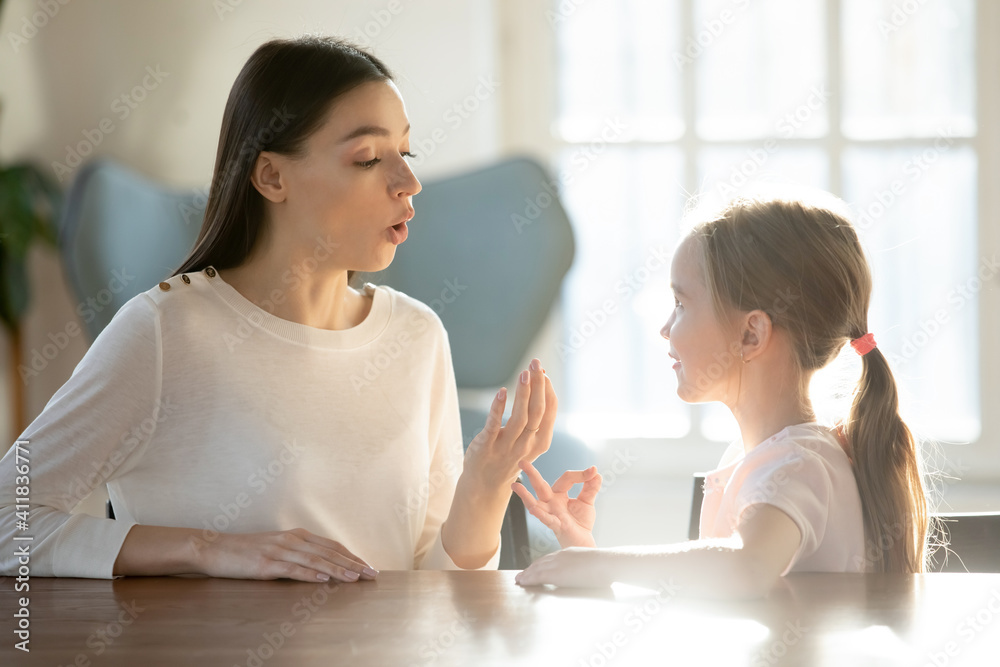 Caring young Caucasian mom talk speak learn pronouncing sounds with small 7s daughter. Millennial female teacher or tutor make hand gesture practice nonverbal sign language with little girl child.