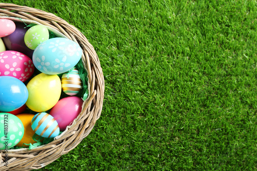 Wicker basket with Easter eggs on green grass, top view. Space for text