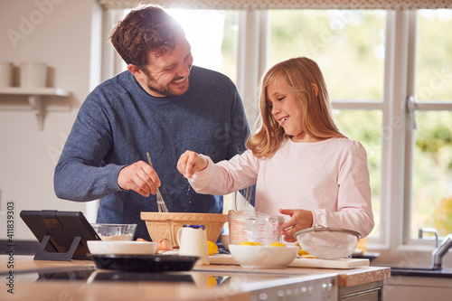 Father And Daughter In Pyjamas Making Pancakes In Kitchen At Home Following Recipe On Digital Tablet