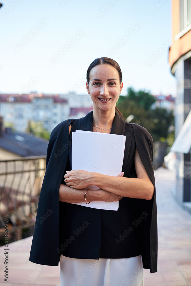 Fototapeta premium Young brunette business woman, wearing stylish black jacket and white dress, holding sheets of paper,standing on balcony. Female portrait in summer. Fashion model on terrace with beautiful city view.