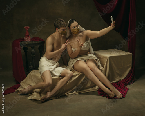 Take selfie for social. Modern remake of classical artwork with modern tech theme - young medieval couple in masks on dark background, golden colored. Concept of technologies, devices, coronavirus, ad