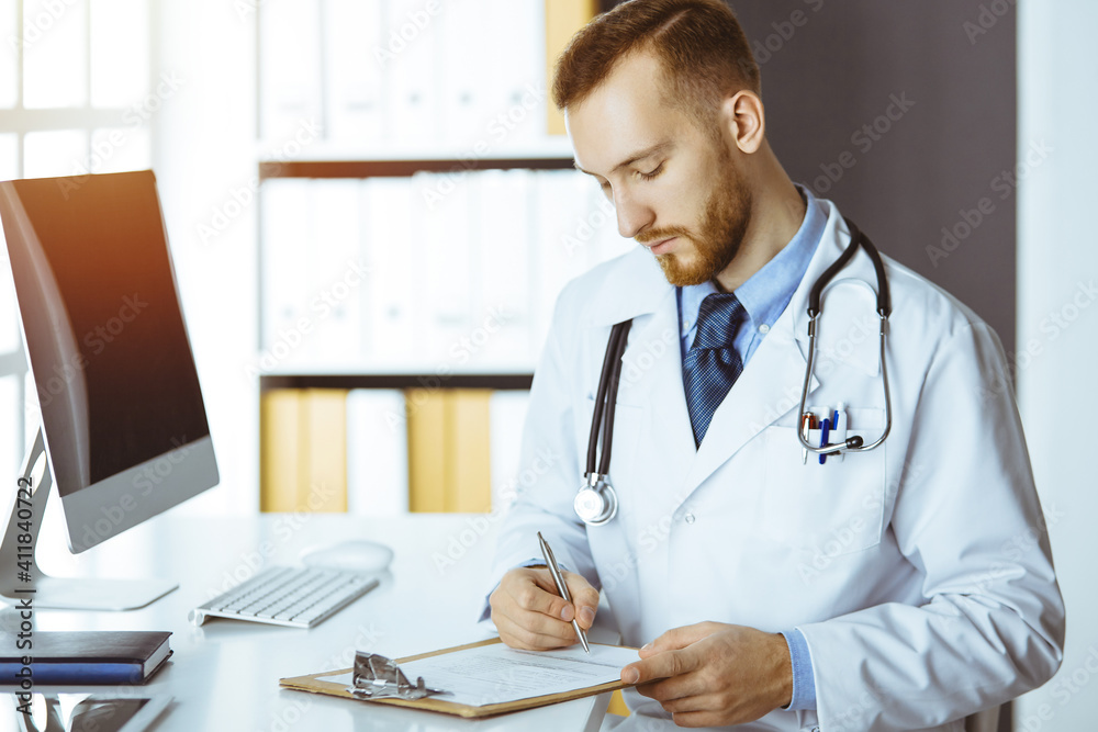 Friendly red-bearded doctor sitting and writing at clipboard in sunny clinic. Medicine concept