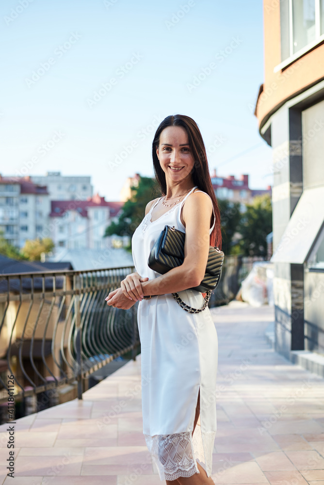Young brunette woman, wearing silk white dress, holding black purse,standing on hotel balcony in the morning. Female portrait in summer. Fashion model posing on terrace with beautiful city view.