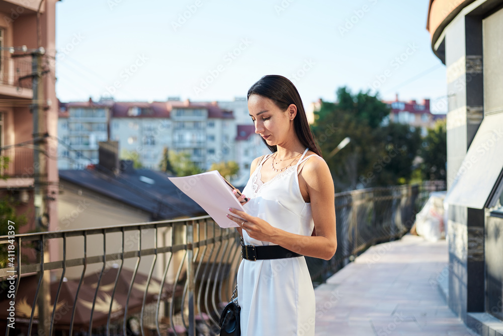 Fototapeta premium Young brunette business woman, wearing silk white dress, holding sheets of paper,standing on hotel balcony. Female portrait in summer. Fashion model posing on terrace with beautiful city view.