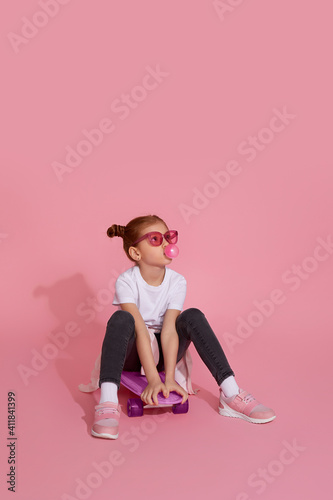 cool redhead little child girl with chewing gum and pink sunglasses sitting on skateboard