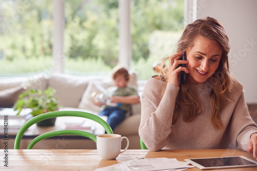 Mother On Phone Call At Home With Digital Tablet Reviewing Domestic Finances With Son In Background photo