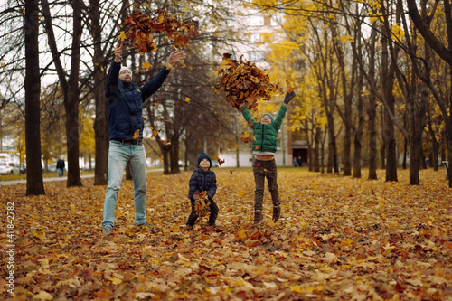 man and two cute caucasian boys throwing yellow fallen leaves and jumping up into the air having fun in autumn park. Father's day concept