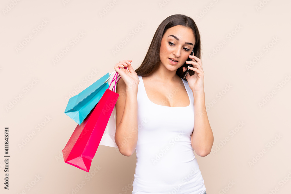 Young caucasian woman isolated on beige background holding shopping bags and calling a friend with her cell phone Young caucasian woman isolated on beige background holding shopping bags and calling a friend with her cell phone