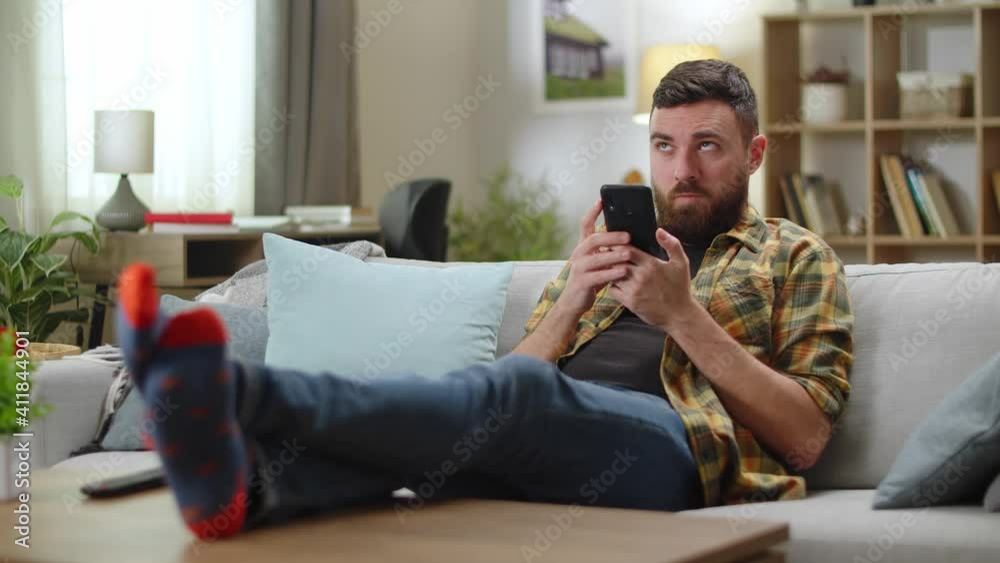 Young man browsing news in mobile phone while sitting on sofa in his living room Young man browsing news in mobile phone while sitting on sofa in his living room