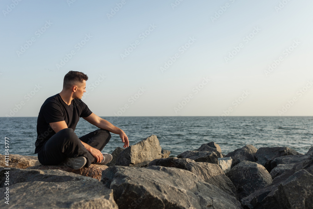 Pensive sports man sits on the coast with stones